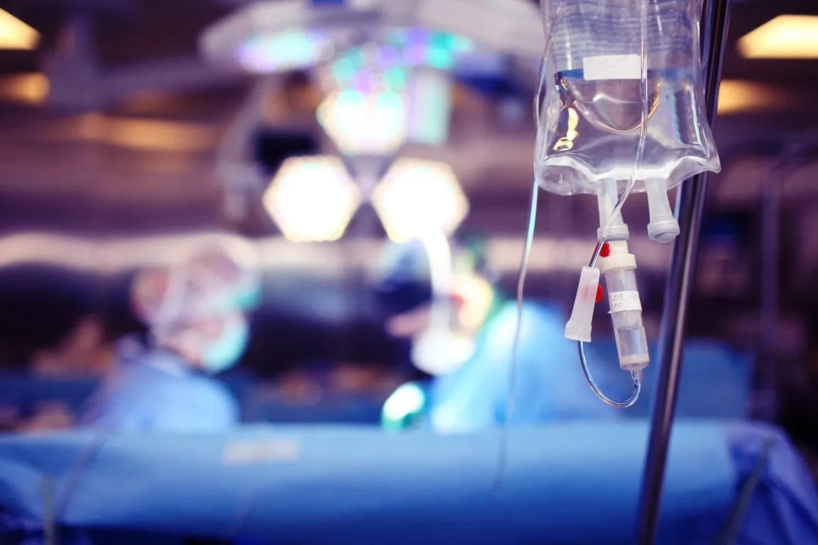 IV Bag in Focus Behind Bed in Operating Room, Two Physicians Working in Blurry Background
