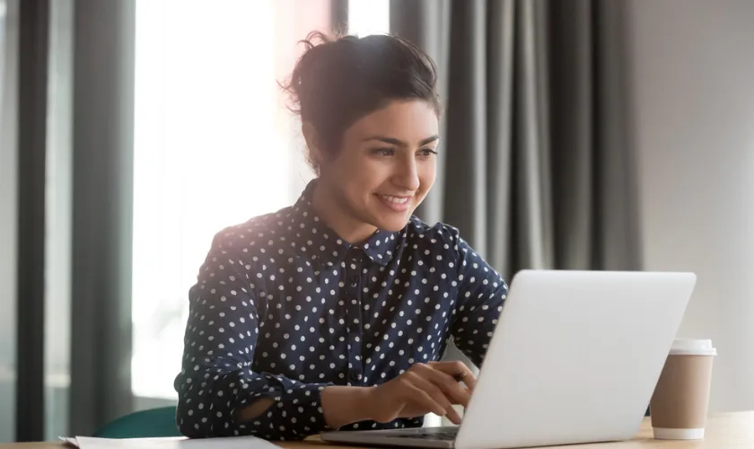 Clinician sits at desk with laptop and coffee_840x500
