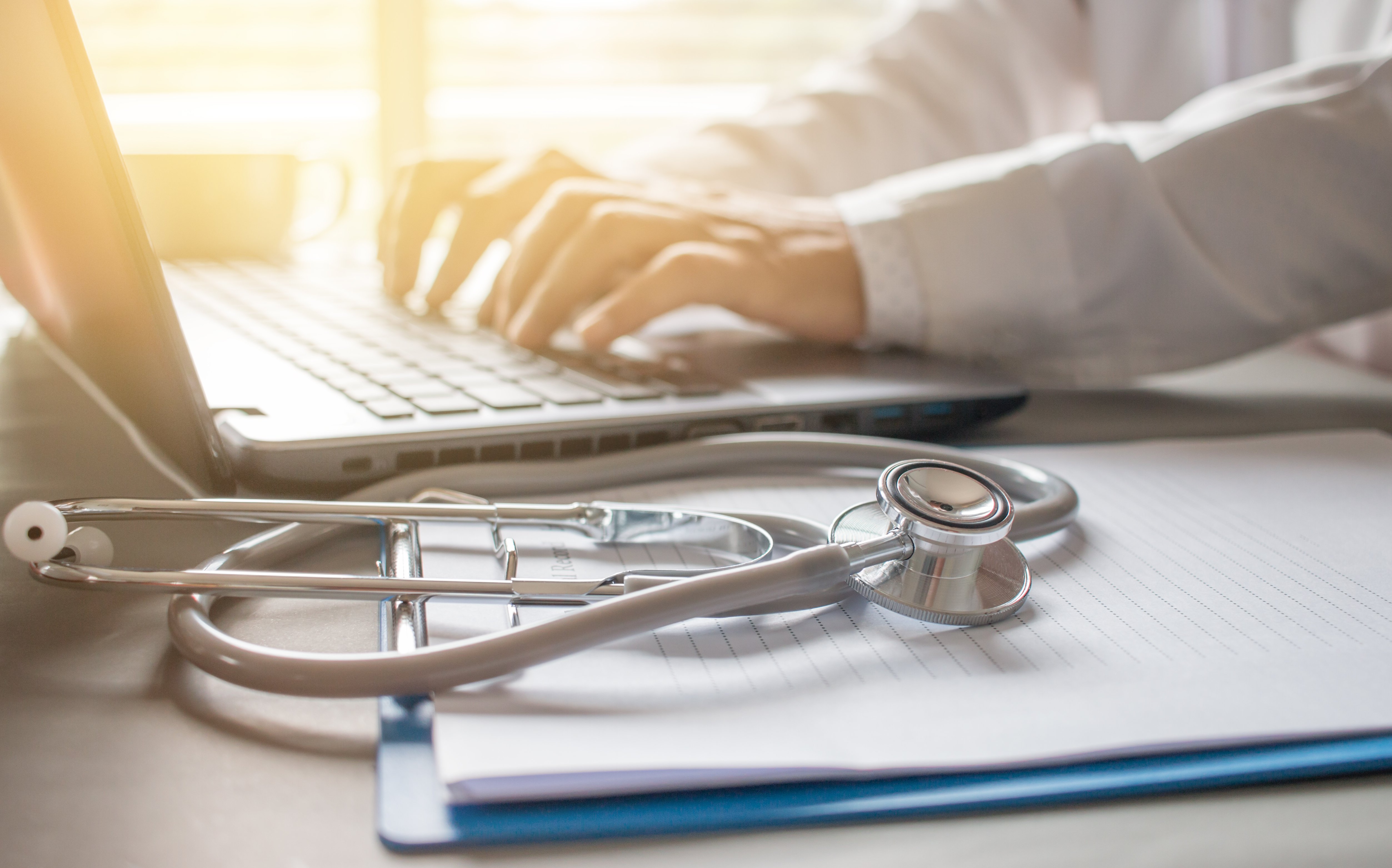 Clinician Working on Laptop with Stethoscope and Notebook on Table