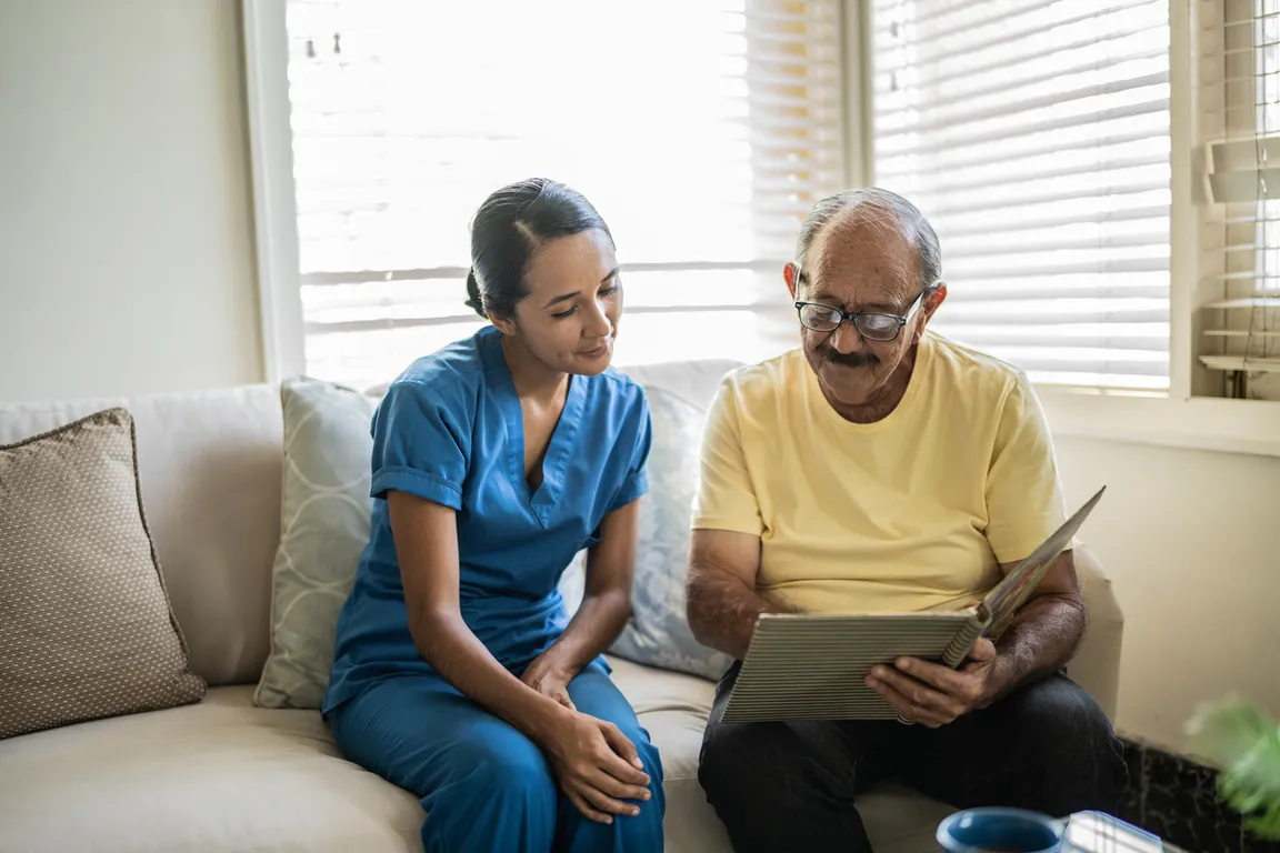 Man shows health care professional family photo album during a home visit; they are both sitting on a couch