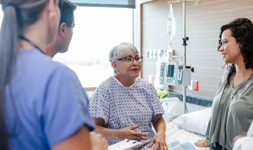 Photo of an older adult patient in a hospital room talking to her health care team and daughter_840x500