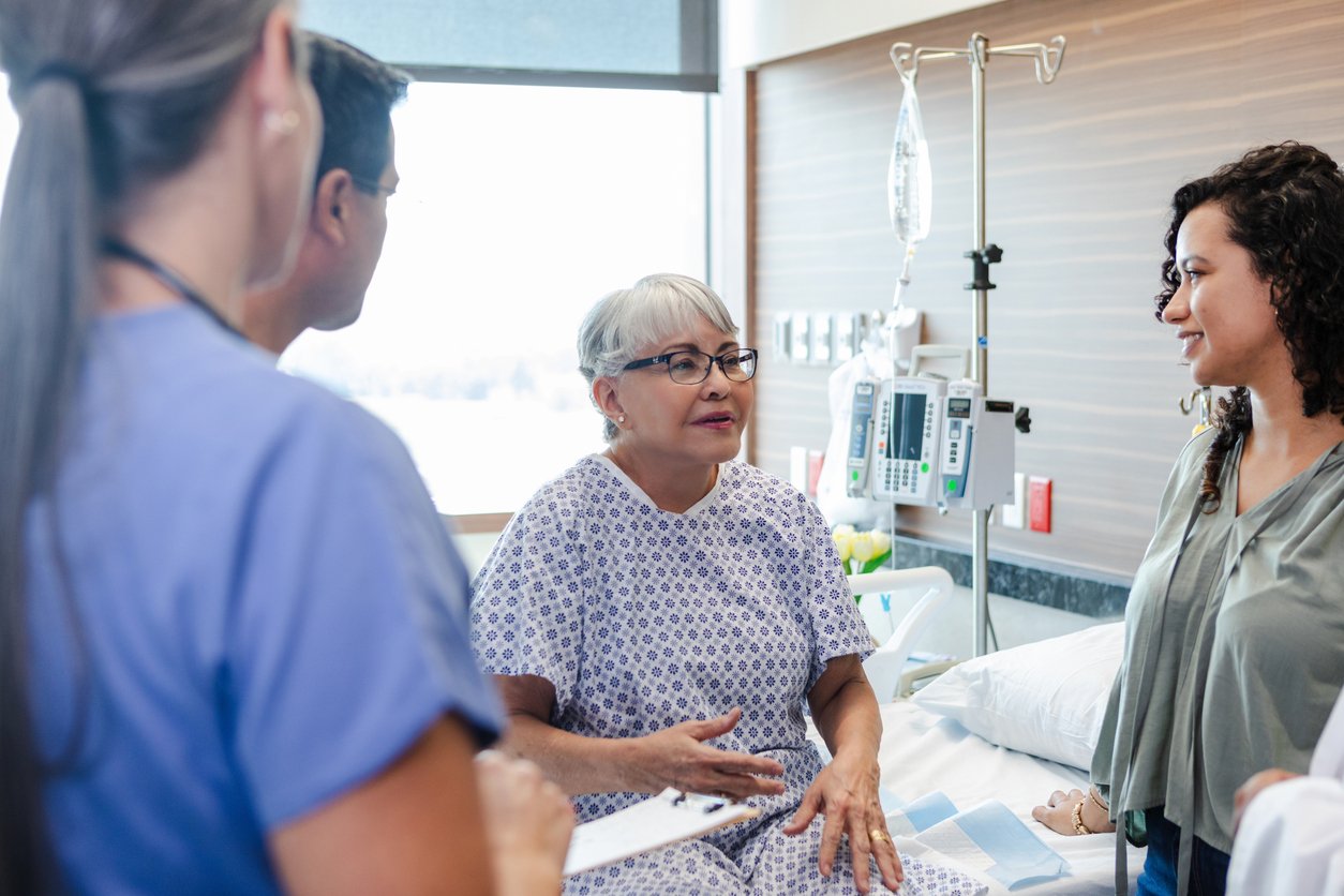 Photo of an older adult patient in a hospital room talking to her health care team and daughter