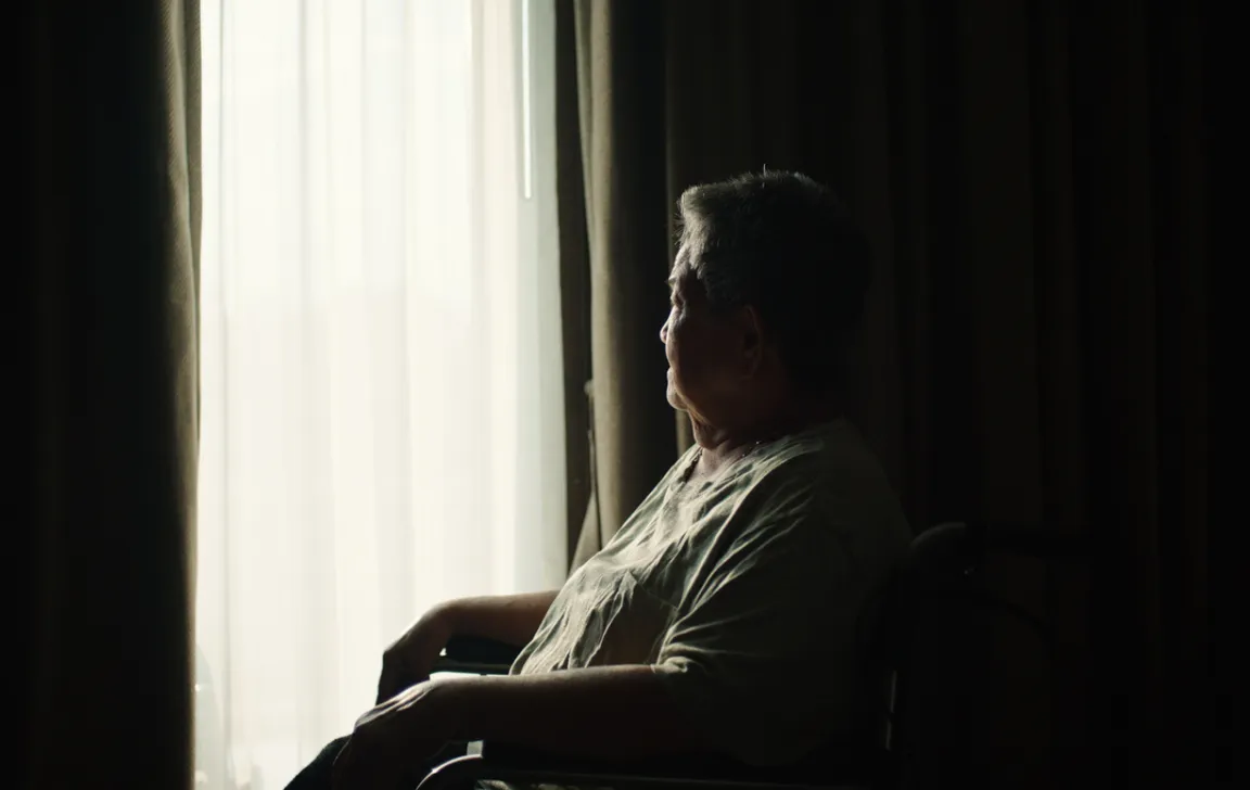 Photograph of an elderly woman sitting in the dark by the window, mostly blending in to the dark background. A panel of light is coming through, showing her silhouette. o show her silouh