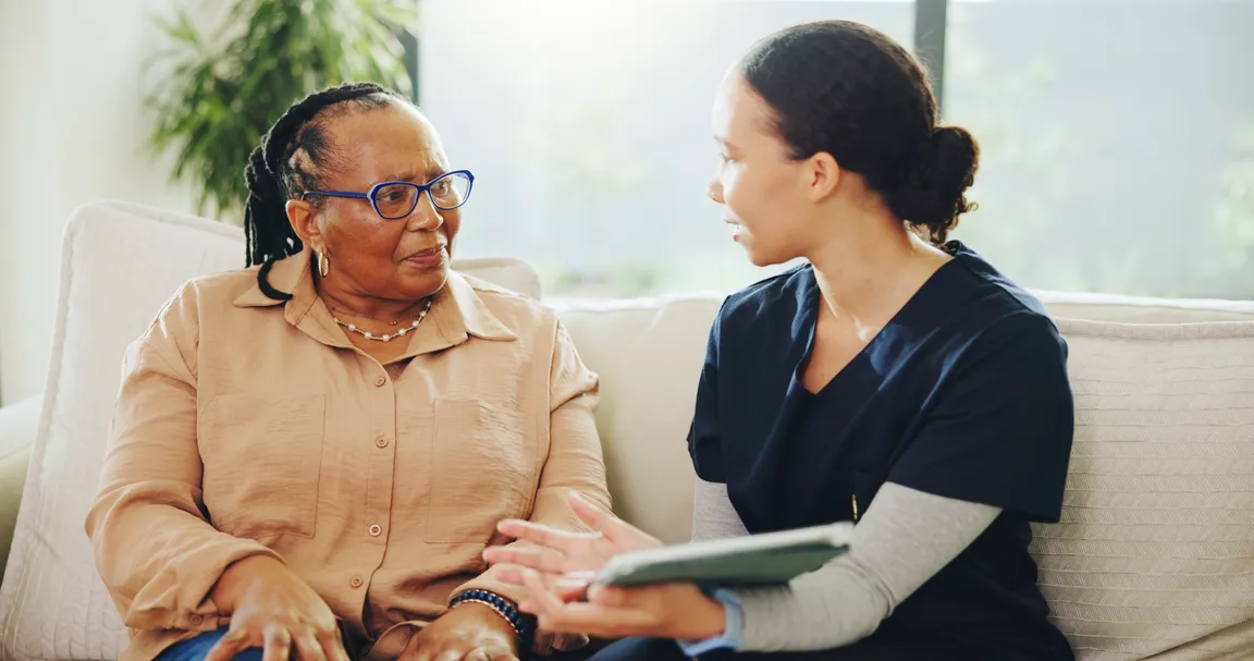 Patient and clinician sitting on couch in a home care setting; clinician holding tablet and talking with patient